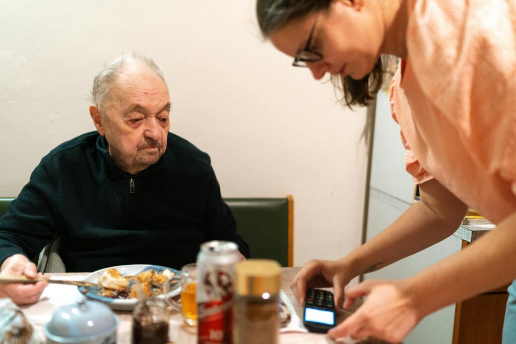 A caregiver assists an elderly man during mealtime at an indoor setting. Captured in Praha.